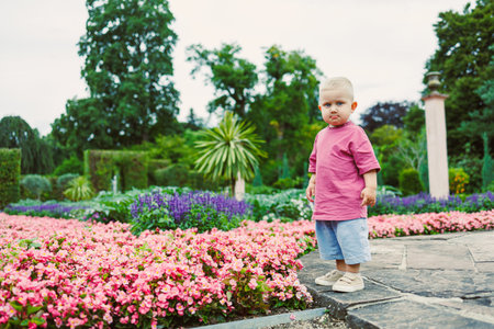 A cheerful toddler stands among vibrant blossoms in a beautiful garden, where lush trees and bright...