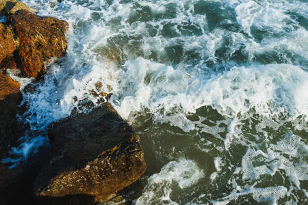 Rugged rocks are partially submerged as ocean waves crash against them, creating white foam under...