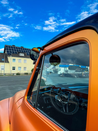 Bright orange classic truck sits parked, showing its shiny interior and reflections of a clear blue sky. Nearby, industrial buildings with solar panels complete the scene.の写真素材