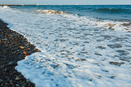 Gentle waves roll onto a rocky beach, forming white foam as they meet the stones under a serene blue sky. The coast stretches far, inviting relaxation and tranquility.の写真素材