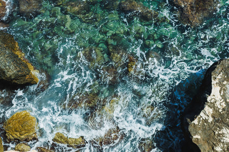 Captured from above, clear turquoise waves crash and foam around rugged rocks. Sunlight glistens on...