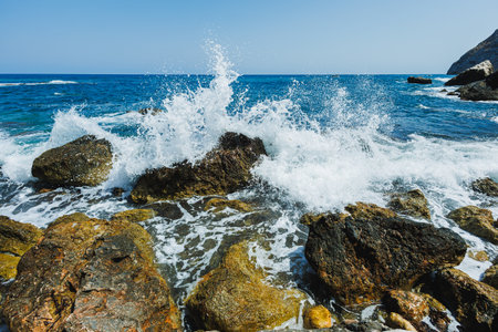 Bright blue waters surge dramatically against weathered rocks, creating frothy splashes under the...