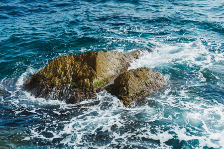 Bright sunlight sparkles on the clear blue ocean as waves roll onto a unique rocky outcrop, creating...