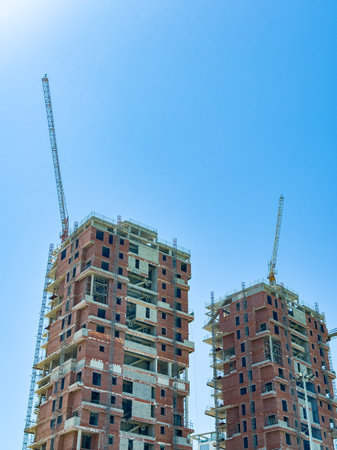 Two tall buildings under construction reach for the sky, surrounded by cranes and workers, showing the vibrant energy of city development on a sunny day.の写真素材