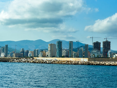 Tall buildings rise along the waterfront, showcasing construction activity. Soft waves lap against the rocky shore beneath a bright sky with clouds and mountains in the background.の写真素材