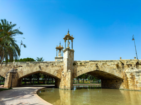 A beautiful stone bridge arches gracefully over still waters, featuring elegant golden towers. Palm trees sway gently in the breeze under the bright blue sky.の写真素材