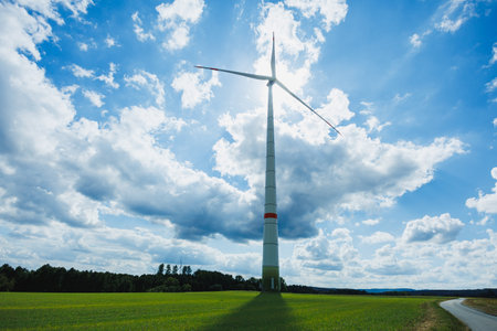 A giant wind turbine captures the gentle breeze, surrounded by lush green fields. Fluffy clouds float across a brilliant blue sky, highlighting the beauty of renewable energy.の写真素材