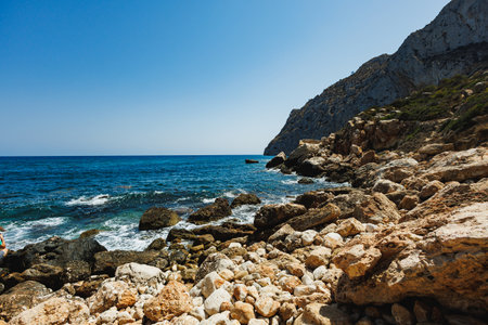 Waves gently crash against the rocky shore while bright sunlight illuminates the vibrant blue water and mountainous terrain, creating a serene coastal atmosphere.の写真素材