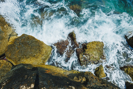 Foamy waves crash against rough, weathered rocks along a vibrant shoreline, reflecting the bright...
