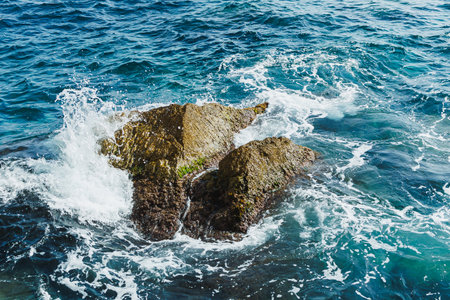 Turquoise water collides with rugged rocks, sending splashes into the air. Bright sunlight reflects...