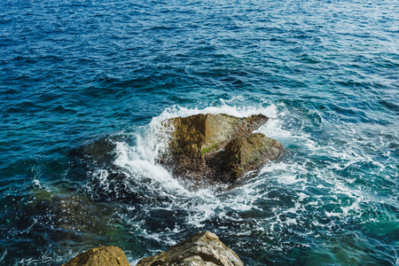 Sunlight glimmers on the surface of the ocean as waves gently hit a rocky outcrop. Sea foam rises...