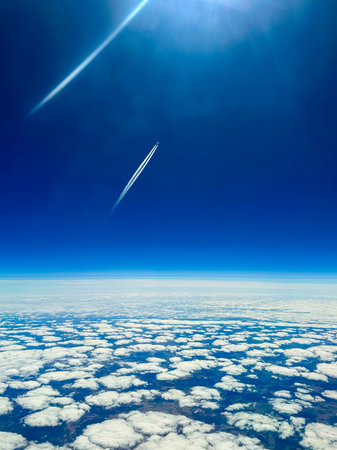 An airplane soars gracefully above the soft, fluffy clouds, tracing a white trail against the vibrant blue sky. The midday light enhances the peaceful scenery.の写真素材