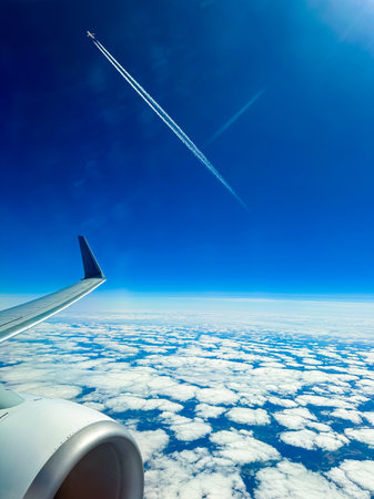 Above the soft white clouds, a jet plane leaves a trail in the bright blue sky while the airplane...