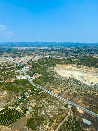 Aerial view showcases a mining area surrounded by greenery and farmland, with a town visible in the background and mountains on the horizon under a clear blue sky.の写真素材