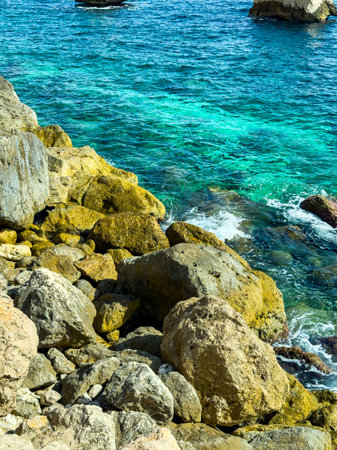 Crystal blue waters gently lap against the golden rocks, creating a peaceful scene along the coastline. A warm sun casts shadows that dance on the water's surface, enhancing the tranquil atmosphere.の写真素材