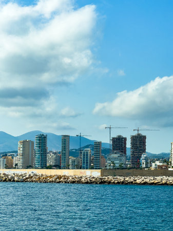 Tall buildings tower above the coastline, showcasing modern architecture against a backdrop of soft clouds and distant mountains. The scene captures the vibrant energy of urban life.の写真素材