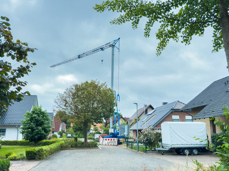 A construction crane stands tall in a quiet residential area, lifting materials amidst houses and trees on a cloudy day. Workers are likely busy on nearby projects.の写真素材