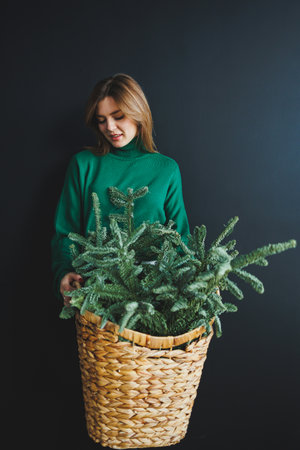 In an inviting moment, a woman in a green sweater tenderly holds a large basket brimming with vibrant evergreen foliage, set against a dark background that highlights the rich textures.の写真素材