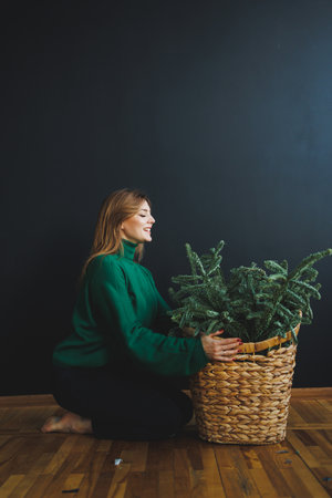 A woman in a green sweater smiles as she lovingly arranges a large potted plant in a woven basket. The cozy indoor setting features a dark wall and warm wooden floors.の写真素材