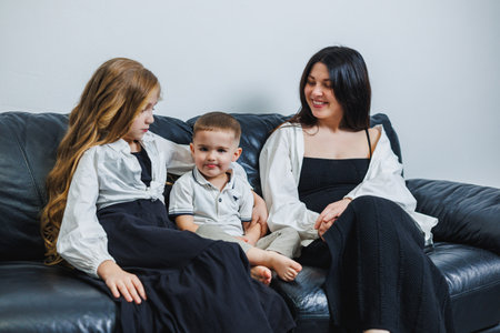 A woman and two children sit together on a dark sofa, sharing laughter and conversation,...