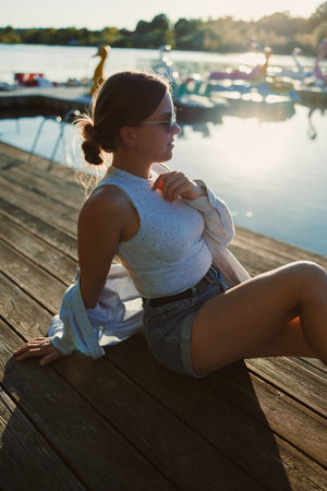 Sitting on a wooden dock by the lake, a young woman in a sleeveless top and shorts basks in the golden glow of sunset. Nearby boats add to the serene atmosphere.の写真素材