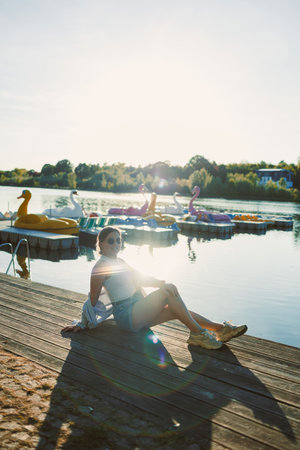 A young woman relaxes by the lake on a sunny day, enjoying the warm weather. Colorful paddle boats float nearby, creating a vibrant scene filled with light and joy.の写真素材