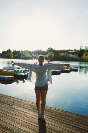 A young woman stands on a wooden dock by a serene lake, arms wide open to welcome the morning sun. Colorful boats float gently in the water, reflecting a peaceful atmosphere.の写真素材