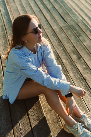 A woman relaxes on a wooden deck, soaking up the sun with a peaceful smile. She wears casual clothing and stylish sunglasses, embodying a laid-back summer atmosphere.の写真素材