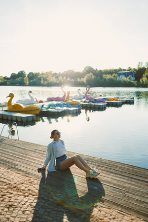 A young woman relaxes on a wooden dock, basking in the sun. Vibrant paddle boats float in the calm lake, surrounded by autumn trees, creating a peaceful and joyful atmosphere.の写真素材