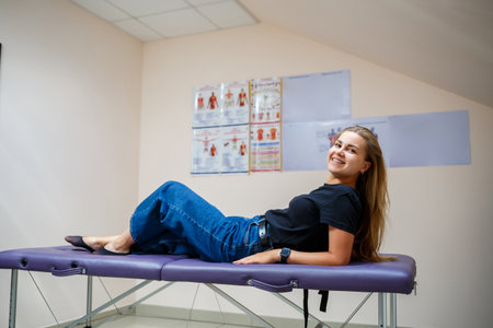 A young woman smiles while reclining on a therapy table in a bright, inviting wellness room. The atmosphere is calm, with informative charts on the walls, suggesting a focus on health.の写真素材