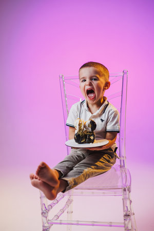 A cheerful young boy sits on a transparent chair with bare feet, excitedly holding a plate of dessert. His laughter fills the colorful room, creating a joyful atmosphere.の写真素材