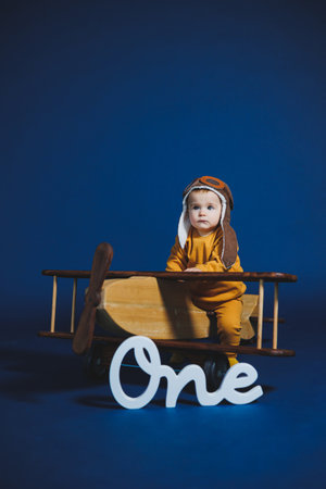 A joyful young child, dressed in a pilot costume and sitting in a wooden airplane, smiles brightly against a deep blue backdrop, marking their first birthday with delight.の写真素材