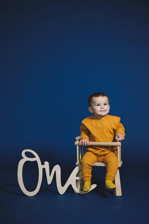 Bright and cheerful setting captures a young child in vibrant yellow sitting confidently on a small chair, next to a sign reading One against a blue backdrop.の写真素材