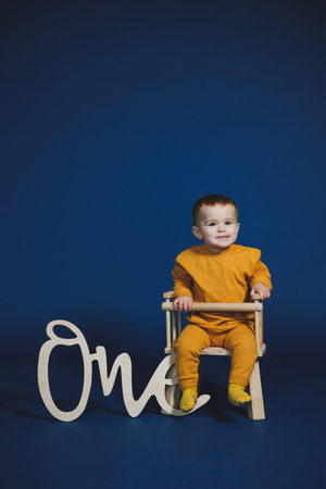 A joyful toddler in a bright yellow outfit sits on a small chair, smiling beside a wooden sign that says one. The vivid blue backdrop enhances the playful atmosphere of the moment.の写真素材