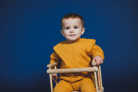 A young child sits confidently in a wooden chair, wearing a cheerful yellow outfit. The background bursts with bright blue, highlighting the child's playful demeanor.の写真素材