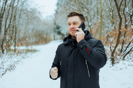 A young man talks on his phone while walking along a snowy path through a serene forest. Trees are...