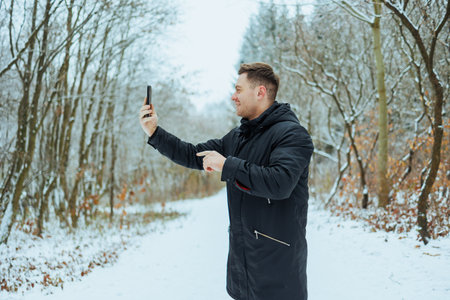 A young man stands on a snow-covered path in a forest, joyfully taking a selfie. The trees are bare...
