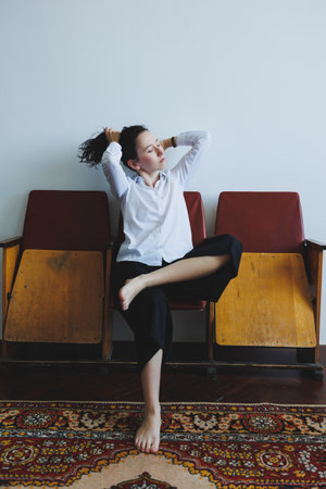 In a serene space, a woman with tousled hair sits on retro chairs. She stretches her arms and...