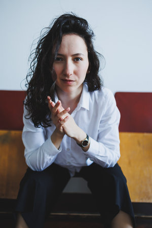 A young woman with curly hair sits confidently on a wooden bench, her hands clasped together as she appears deep in thought in a cozy, softly lit room.の写真素材