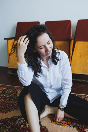 A young woman with curly hair smiles while gently sitting on a vibrant rug, surrounded by warm wooden chairs, enjoying a peaceful afternoon in a sunlit space.の写真素材