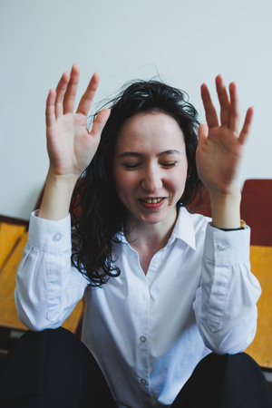 A young woman with curly hair smiles brightly, raising her arms in joy. The relaxed indoor setting...