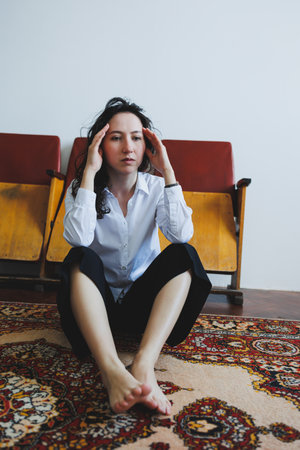 A woman sits cross-legged on a vibrant carpet, fingers pressed to her temples. Her expression shows deep contemplation, surrounded by simple decor and vintage chairs in a quiet space.の写真素材