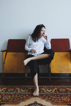 A young woman sits pensively on a vintage chair, dressed casually in a white shirt and dark pants, surrounded by a warm ambiance that inspires creativity and reflection.の写真素材