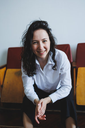 A young woman with curly dark hair smiles warmly while sitting on retro seats in a bright, inviting...