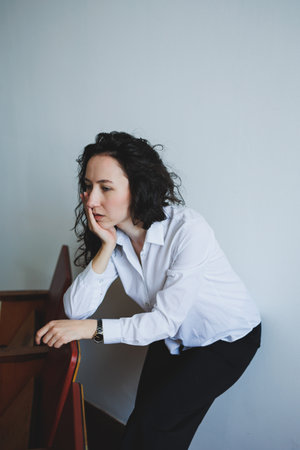 A woman with curly hair in a white shirt rests her chin on her hand while leaning against a piece of furniture in a simple, bright room, engaged in deep thought.の写真素材