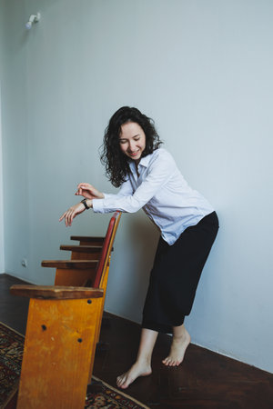 With a joyful expression, a woman twirls barefoot near a wooden chair, embracing the bright and airy ambiance of a simple, sunlit room filled with warmth.の写真素材