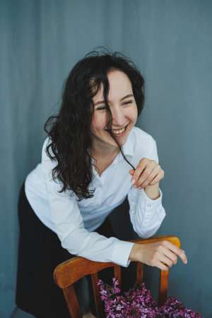 In a calm, minimalist space, a young woman with curly hair smiles widely as she playfully touches...