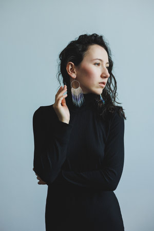 A woman with curly hair poses gracefully in a black top, showing her unique earrings. The soft blue background emphasizes her elegance and style, creating a serene atmosphere.の写真素材