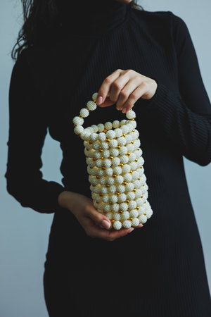 A person stands in a softly lit studio, showcasing an elegant handmade pearl bag adorned with golden beads. They wear a sleek black dress, enhancing the bag's charm and sophistication.の写真素材