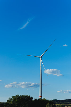 A large wind turbine rises above a green landscape, its blades turning gently in the wind. White...